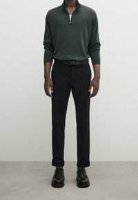 Pull à fermeture éclair col montant vert foncé porté sur une chemise blanche, associé à un pantalon noir fuselé et des chaussures noires à lacets, créant un look élégant.