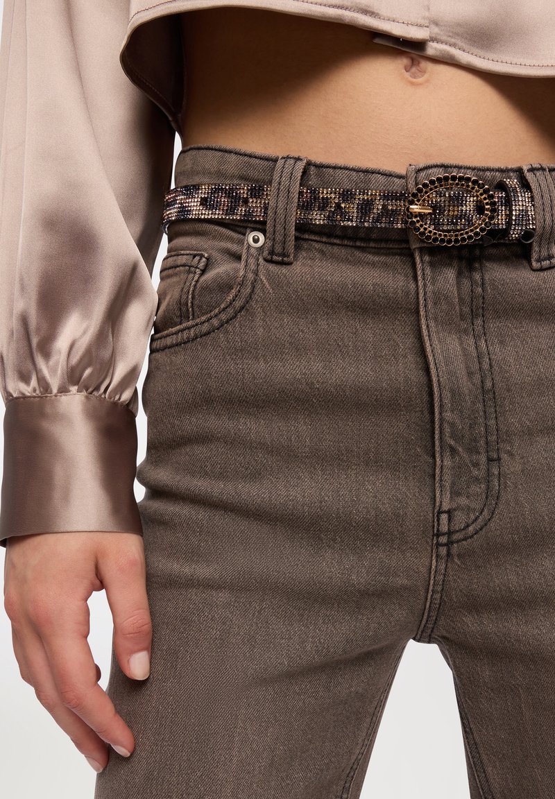 Jeans in denim marrone con dettagli di texture, abbinati a un top lungo in satin beige lucido a maniche lunghe e una fibbia per cintura ovale adornata di gioielli.