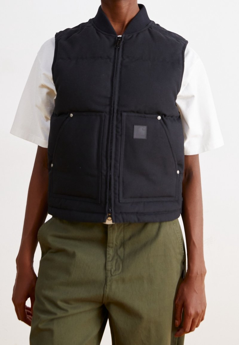 Černá vycpaná vesta se zapínáním na zip, vybavená dvěma velkými kapsami, texturovaným povrchem a malou nášivkou s logem. Nositelná přes bílou košili.