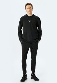 Sudadera deportiva negra con bolsillo frontal y texto bordado, combinada con joggers negros ajustados. La tela parece suave y elástica.