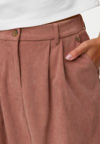 Pantalons en velours côtelé marron avec des plis sur le devant, une fermeture à bouton marron et des poches latérales. Le tissu a une texture douce et côtelée.