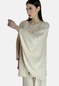 Top beige, dalla vestibilità ampia con maniche larghe, scollo rotondo e texture liscia; il tessuto leggero si drappeggia dolcemente; indossato con pantaloni abbinati.