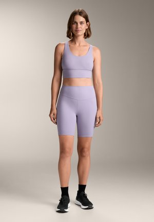 Svetlofialový športový crop top a zodpovedajúce šortky s vysokým pásom z elastického materiálu, doplnené o čierne tenisky a členkové ponožky.