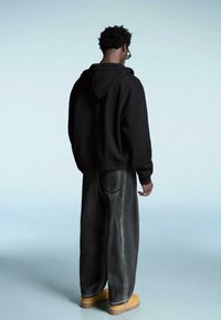 Černá mikina s kapucí, volné tmavé džíny a žluté dřeváky. Jednoduchý design s viditelnými švy a uvolněným střihem.