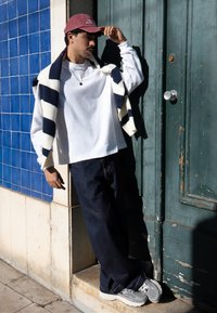 Jonge man in een witte casual trui, brede marineblauwe broek, grijze sneakers, gestreepte trui over de schouders gedrapeerd, verstelend aan een bordeauxrode pet bij een blauwe betegelde muur.