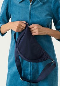 Borsa a spalla a forma di mezzaluna di colore blu navy, realizzata in tessuto testurizzato e dotata di una tracolla regolabile, tenuta da una persona che indossa un completo in corduroy blu.