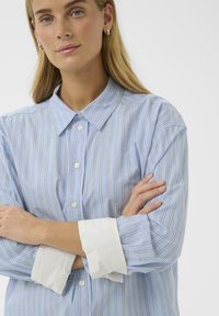 Chemise bleu clair à boutons avec des rayures verticales blanches et dorées, dotée d'un col pointu et de poignets blancs contrastants. Texture de tissu lisse.