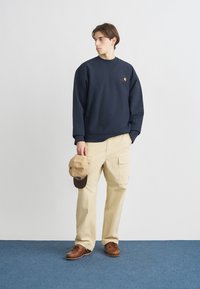 Ναυτικό φούτερ με λογότυπο, συνδυασμένο με μπεζ παντελόνι cargo και καφέ παπούτσια. Ένα καφέ καπέλο με σκουρόχρωμη γείσο κρατείται στο αριστερό χέρι.