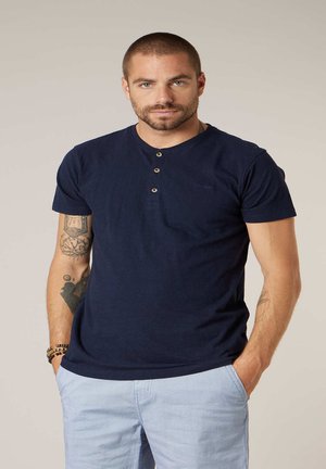 Mann mit kurzen Haaren und Tätowierungen trägt ein marineblaues Henley-Shirt und hellblaue, gestreifte Hosen, steht mit den Händen in den Taschen.