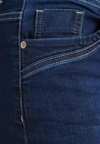 Vaqueros de mezclilla en azul oscuro con un acabado texturizado. Presenta un pequeño bolsillo frontal, costuras blancas contrastantes y un detalle de botón de metal.