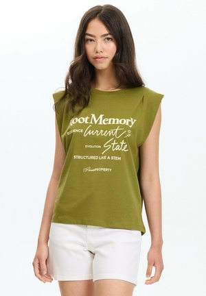 Joven con cabello largo y oscuro, lleva una camiseta sin mangas verde oliva con texto blanco y pantalones cortos blancos, de pie frente a un fondo liso.