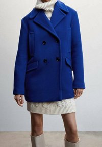 Blauwe wollen peacoat met een dubbelzijdige ontwerp, zwarte knopen, zijzakken en een uitgesneden kraag. Draagt over een crèmekleurige gebreide jurk.
