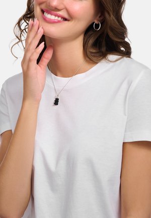 Weißes Baumwoll-T-Shirt mit kurzen Ärmeln. Silberne Kette mit einem kleinen schwarzen Bärenanhänger. Das Modell berührt sanft ihr Gesicht.