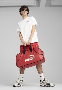 Rode duffeltas met zwarte handvatten, met een opvallend wit Puma-logo. Model draagt een wit shirt en rode geblokte shorts.