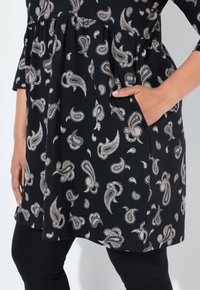 Schwarzes Kleid mit beigem Paisleymuster, Hand in der Seitentasche, kombiniert mit schwarzen Leggings, die bis zur Mitte des Oberschenkels und bis zum Handgelenk und zur Hand sichtbar sind.