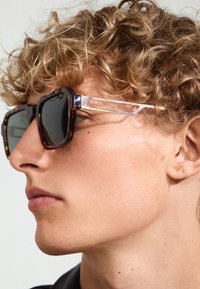 Lunettes de soleil en écaille de tortue avec des montures carrées, des verres sombres et des accents métalliques sur lestempes. Design texturé et embouts d'oreille transparents.