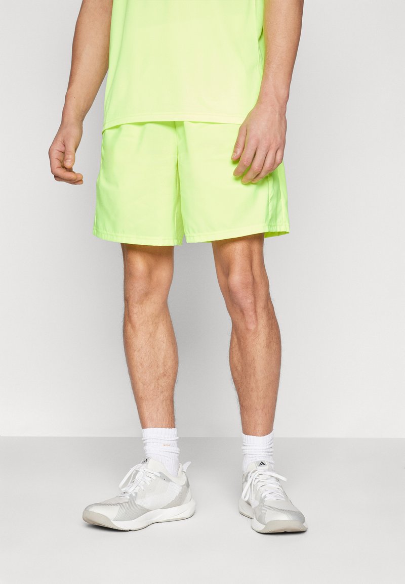 Neon-gelbe Sportshorts mit einem elastischen Bund, leichtem Material und lockerer Passform. Getragen mit weißen Sportschuhen und Socken.