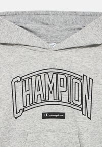 Γκρι φούτερ με κουκούλα, με μεγάλο μαύρο γράφημα "CHAMPION" και μια μικρή μαύρη ετικέτα με το όνομα της μάρκας. Μαλακή υφή υφάσματος.