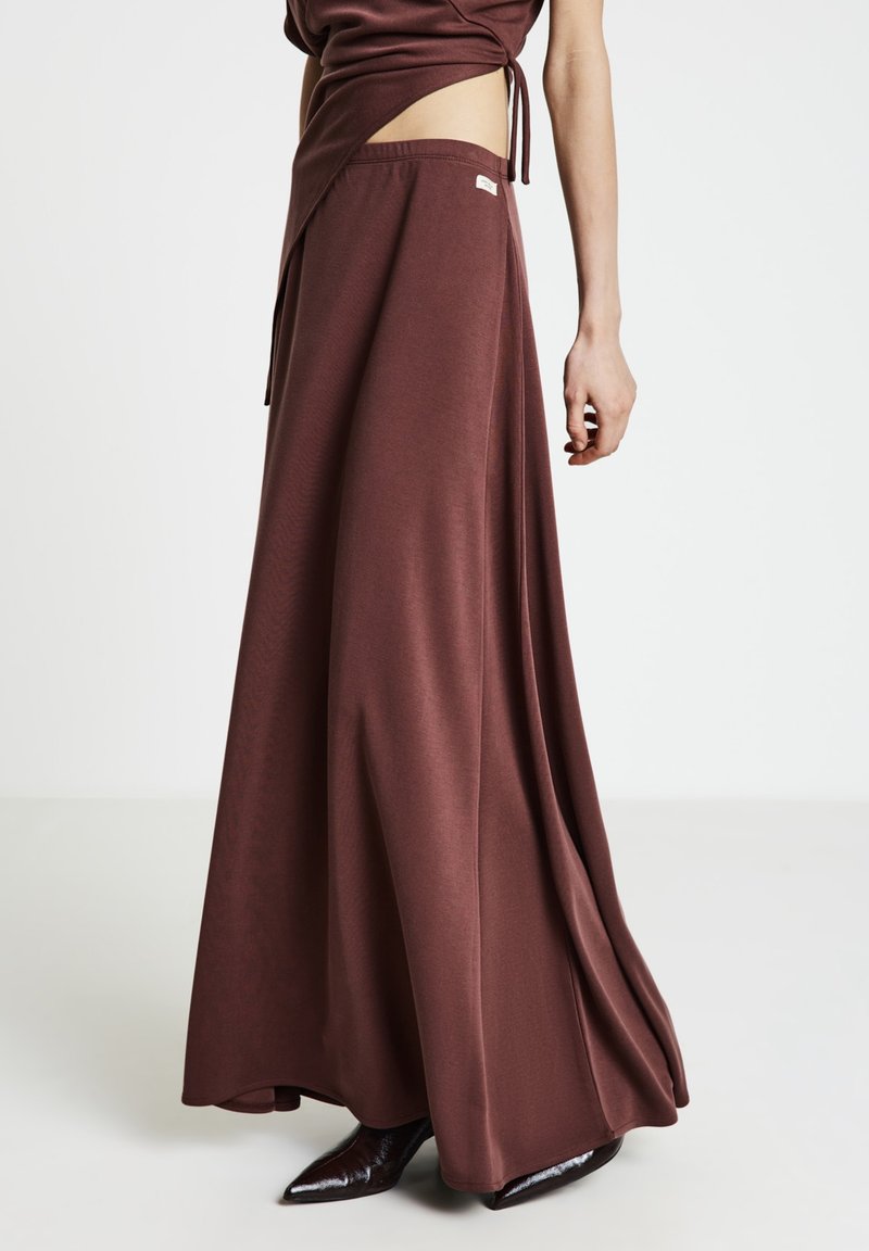 Maxirock aus tiefbraunem Stoff mit breitem Bund, weichem Fall und dezentem Logodetail; kombiniert mit spitzen schwarzen Stiefeletten.