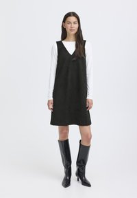 Schwarzes Wildleder-Rippkleid mit V-Ausschnitt und ausgestellt Silhouette, über einem weißen Langarmshirt getragen, kombiniert mit schwarzen kniehohen Stiefeln.