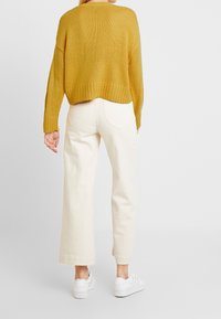 Žlutý pletený svetr s krátkým střihem, doplněný o široké krémové kalhoty a bílé tenisky, zobrazený zezadu.