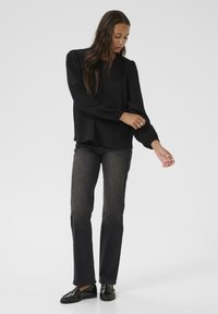 Blouse noire à manches longues en tissu texturé, coupe décontractée et poignets élastiques, assortie à un pantalon en denim foncé et des mocassins noirs.