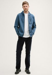 Niebieska bluza z kapturem, z kieszenią z przodu i kieszenią na zamek na klatce piersiowej, noszona na białej koszulce, w połączeniu z ciemnymi spodniami i czarnymi butami.