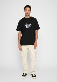 Sort bomulds t-shirt med et grafisk motiv af en fugl i flugt, parret med cremefarvede cargo bukser og sorte sneakers med hvide accenter.