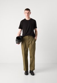 Black short-sleeve t-shirt, olive green trousers with pleated front, worn with black boots, and holding a black bag. Simple, functional design.