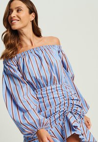Blau gestreiftes Off-Shoulder-Kleid mit langen Ärmeln, gerafftem Bund und fransigem Saum. Mit abwechselnd breiten braunen und schmalen weißen Streifen.
