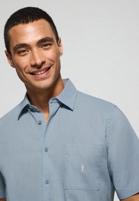 Camicia a maniche corte di colore blu chiaro in un tessuto morbido, con chiusura a bottoni, taschino sul petto e un piccolo dettaglio ricamato.