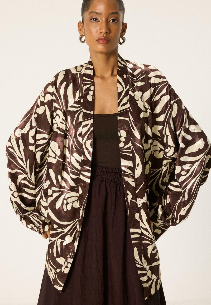 Femme portant une veste oversize marron et beige à motif floral par-dessus un haut noir et une jupe foncée, les mains dans les poches.