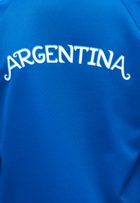 Μπλε ύφασμα με την λέξη "ARGENTINA" τυπωμένη με λευκά γράμματα, κυρτά πάνω από το πάνω μέρος του στήθους.