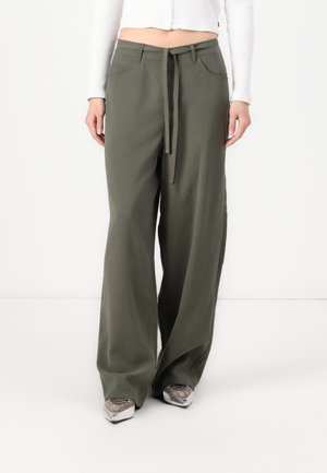 Persoon die losse donkergroene broek met wijde pijpen en tunnelkoord taille draagt, met een wit geribd cropped shirt met lange mouwen en zilveren puntige schoenen.