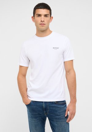 Weißes Baumwoll-T-Shirt mit kurzen Ärmeln, rundem Halsausschnitt und einem kleinen schwarzen „MUSTANG“-Logo auf der Brust. Kombiniert mit blauen Denim-Jeans.