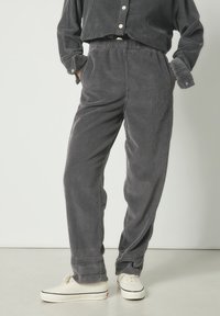 Personne portant un pantalon en velours côtelé gris foncé et une chemise assortie avec des baskets blanches, debout les mains dans les poches devant un fond uni.