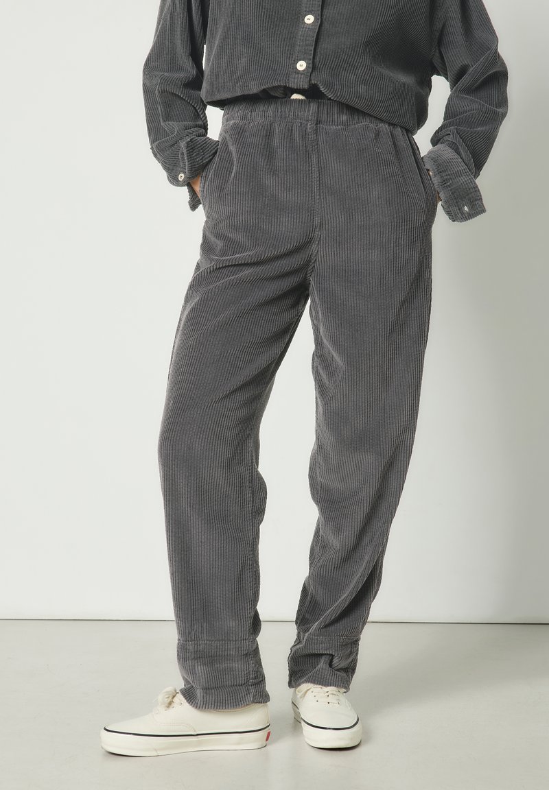 Personne portant un pantalon en velours côtelé gris foncé et une chemise assortie avec des baskets blanches, debout les mains dans les poches devant un fond uni.