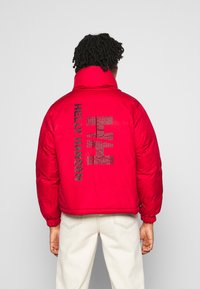 Rød pufferjakke med høj krave, elastiske manchetter og et stort sort "HELly HANSEN" logo på ryggen med lodrette striber.