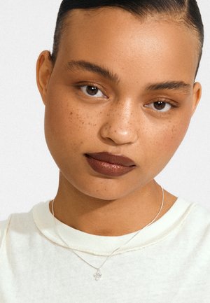 Sølv halskæde med vedhæng båret over en lysekræmd T-shirt. Personen har en glat hudtone, brune øjne og mørke læber.