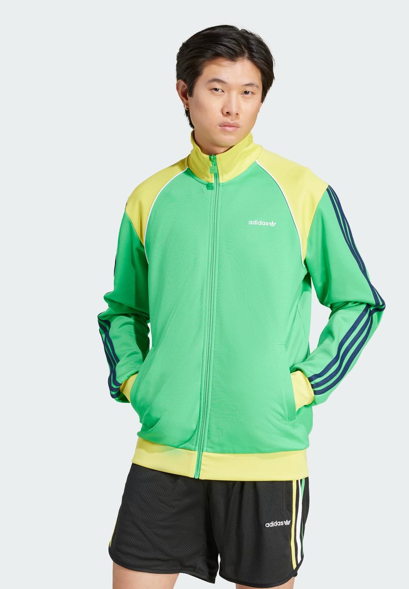 Zelena zip-up trenirka s rumenimi detajli, ki ima visok ovratnik, bel Adidasov logo in mornarsko modre črte po rokavih.