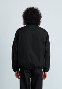 Casaco preto de impermeável com um ajuste descontraído, punhos e bainha elásticos. Tecido suave, design minimalista e gola alta, mostrado por trás.