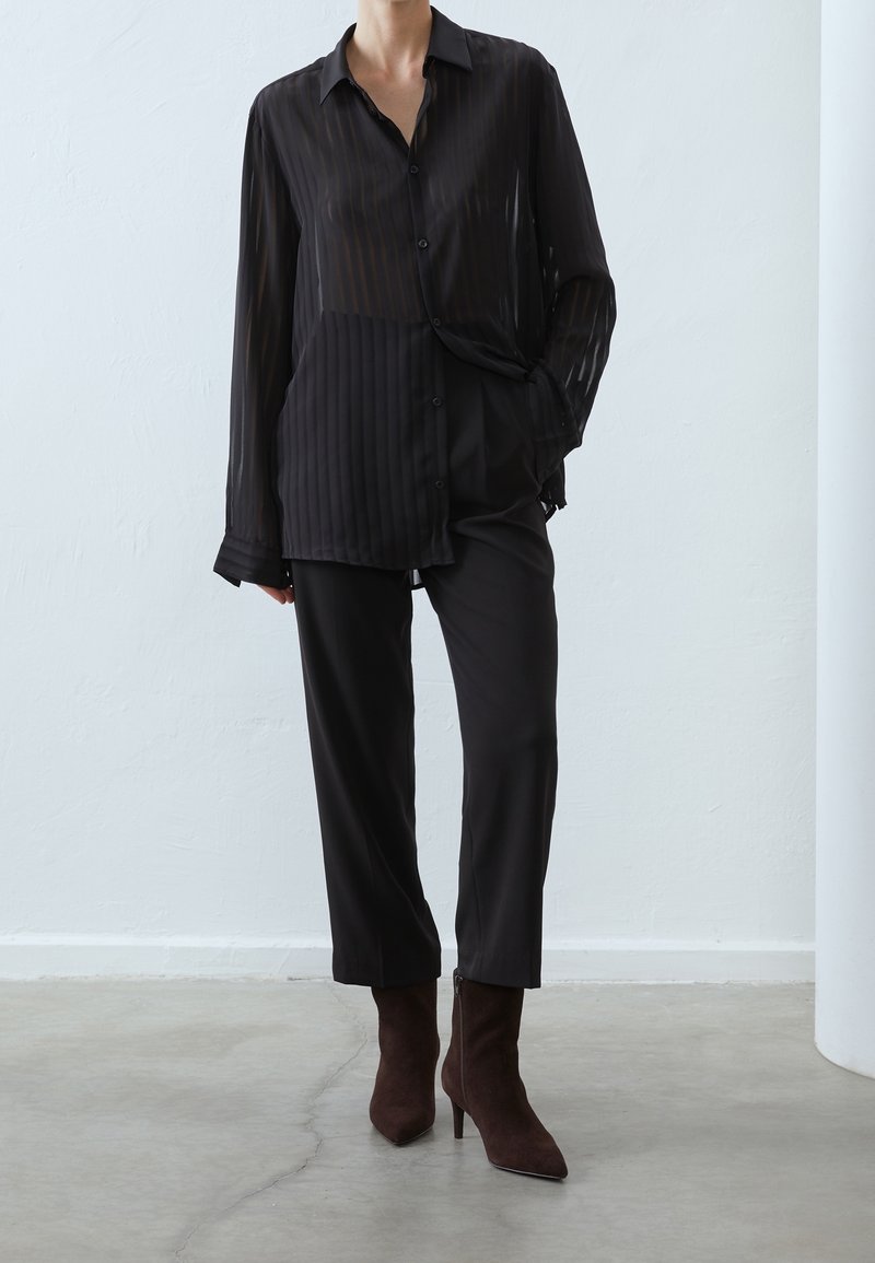 Personne portant une chemise transparente noire à rayures avec boutons, un pantalon noir court et des bottines à talons brun foncé, debout à l'intérieur.