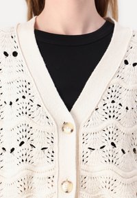 Ljusbeige virkad cardigan med v-ringning, blommönster och två runda knappar fram. Bärs över en svart ribbad topp.