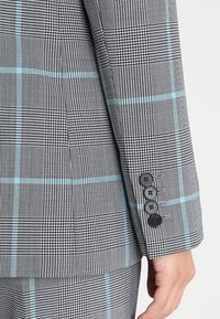 Grå rutig kostymärm med blå linjer. Har tre svarta knappar och textil med varierande mönstertäthet.