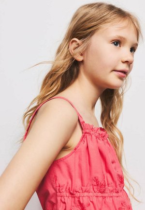 Jeune fille aux longs cheveux blonds portant une robe rose sans manches, regardant pensivement vers la droite sur un fond uni.