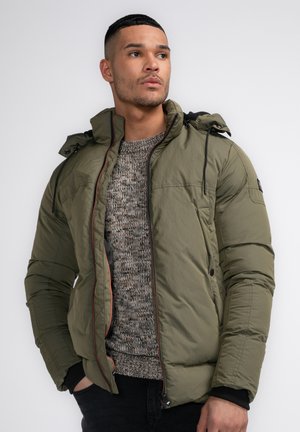 Doudoune vert olive avec capuche, détails noirs, fermeture éclair avant et poignets élastiques, portée sur un pull gris texturé.