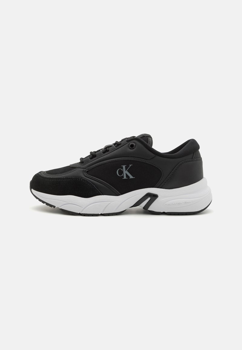 Črne športne čevlje z mešanico gladkih in semiš tekstur. Imajo belo podplat, zaobljeno obliko in logotip v srebrni barvi na strani.