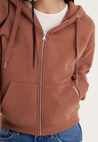 Sudadera con capucha color óxido con cremallera, hecha de tela suave, con capucha forrada, bolsillos tipo canguro y puños acanalados. Presenta herrajes de cremallera plateados.