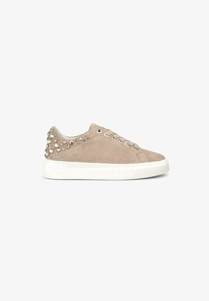 Beige suède sneaker met een witte rubberen zool, ronde neus en metalen studs aan de buitenkant, voorzien van een standaard vetersluiting.