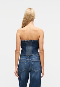 Ξανθιά γυναίκα που κοιτάζει προς τα πίσω, φοράει κορυφή από τζιν χωρίς τιράντες και ψηλόμεσα τζιν παντελόνια, με φόντο ένα λευκό απλό φόντο.